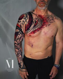 tatuaje en el pecho y tatuaje en el brazo en negro y rojo hecho por abel miranda. el tatuaje del pecho cruza en diagonal desde el pectoral al hombro opuesto.